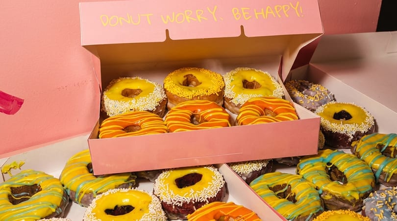 Pink boxes overflow with colorful donuts, some with yellow, orange, or blue icing and sprinkles.