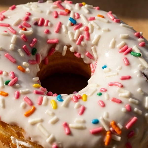 A close-up of a white-frosted donut with colorful sprinkles on a brown surface.