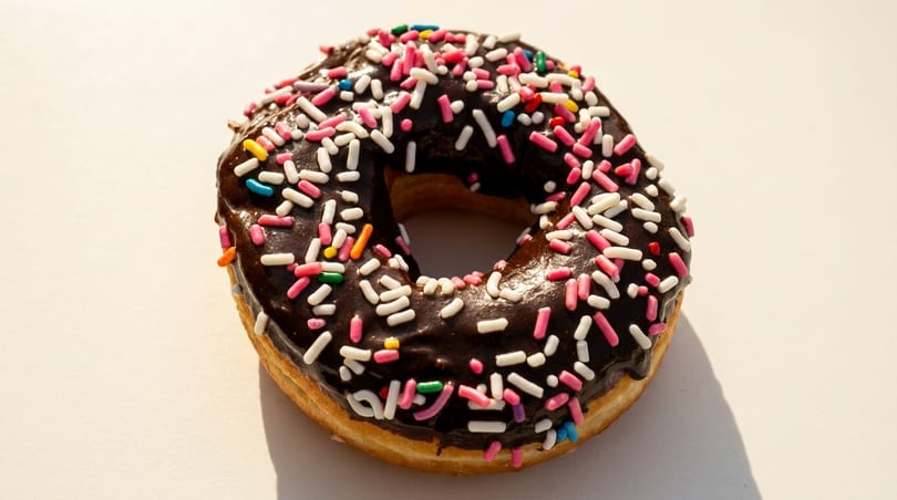 A chocolate-frosted donut with colorful sprinkles sits on a white surface.