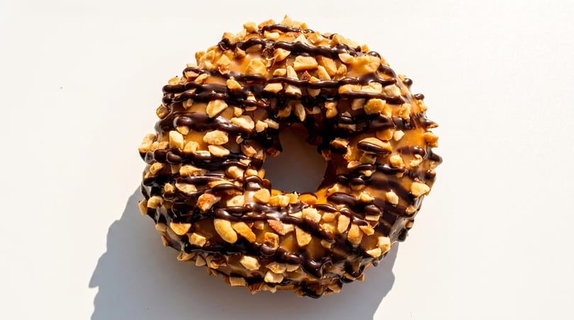 A peanut butter donut with chocolate drizzle and chopped peanuts on a white surface.