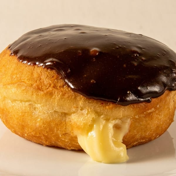 A Boston cream donut with chocolate icing and vanilla cream oozing out, on a white plate.