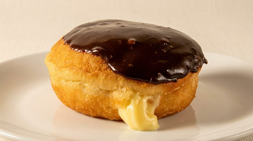 A Boston cream donut with chocolate icing and vanilla custard oozing out, on a white plate.